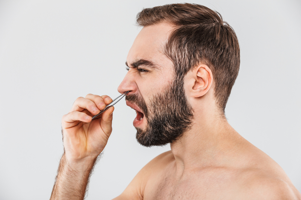 Close up portrait of a suffering bearded man standing isolated over white background, plucking out his nose hair