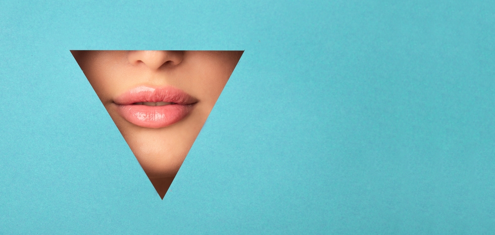 A woman face is visible through a geometric cutout in the shape of a triangle. She is looking directly at the camera, with one eye partially covered. The background is a simple, blank surface.